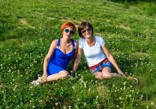Two young beautiful sisters with sunglasses sitting on the grass and sincerely smiling on a summer day in Russia