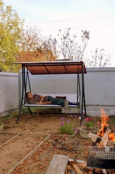 Sleeping young man laying on a garden swing next to bonfire
