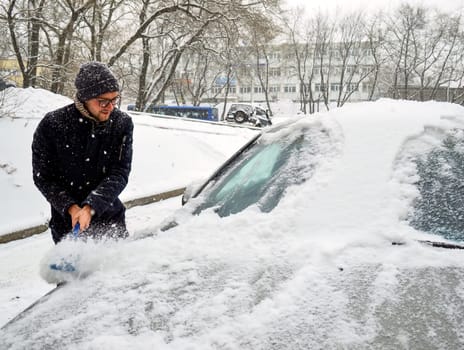 Young man sweeps snow from the car on a snowy day in winter