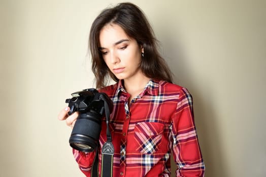 Tired serious woman photographer in red shirt holding camera and looking at bad,failed and unsuccessful photos on beige background. Upset about the result. Copy space