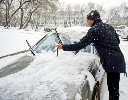 Young man sweeps snow from the car on a snowy day in winter