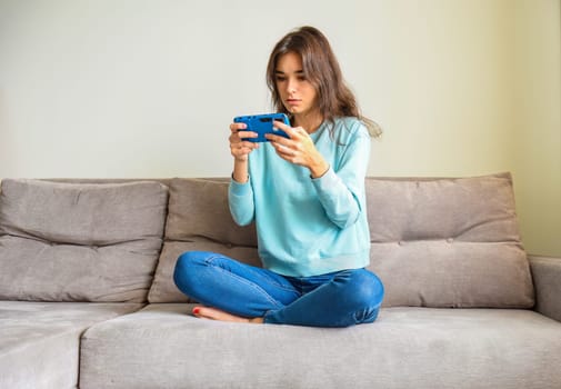 Surprised young woman holding smartphone with two hands,sitting on the sofa in casual clothes. She reading new or playing games. Lockdown activities