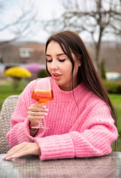 Vertical portrait of young russian woman drinking cocktail aperol spritz, sitting outdoors in pink sweater