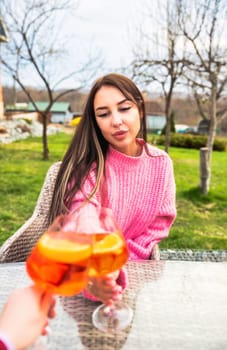 Portrait of young russian woman drinking cocktail aperol spritz, sitting outdoors in pink sweater