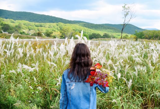 Young woman holding bouquet of autumn wildflowers and branches with red leaves on her shoulder, standing with her back to the camera and looking at field of Miscanthus sinensis (Chinese silver grass).