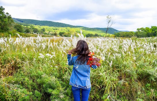 Young woman holding bouquet of autumn wildflowers and branches with red leaves on her shoulder, standing with her back to the camera and looking at field of Miscanthus sinensis (Chinese silver grass).