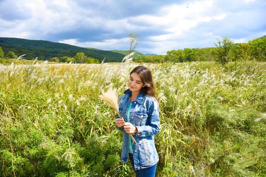 Young smiling woman holding bouquet of Miscanthus sinensis (Chinese silver grass) in her hands and looking at it against the background of a field. Autumn in Russia. Copy space