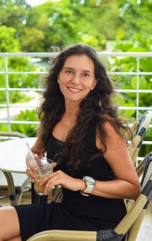Portrait of 40 years old russian woman without make up sitting with glass of ice coffee in black dress