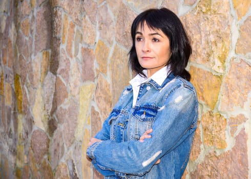 Beautiful Russian 45 y.o. woman in denim jacket looking away with arms crossed and standing against the wall. She is serious and confident