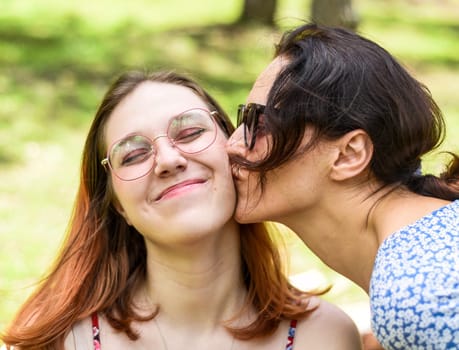Mother kissing her teenage daughter with dyed hair and glasses against of green leaves