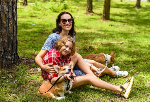 Mother hugging her little daughter resting on her sholder with closed eyes, with two corgi dogs against of green grass in the forest