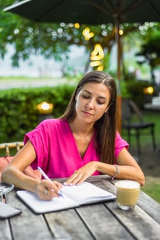 Cute woman sitting on a chair in summer garden and writing in a notebook