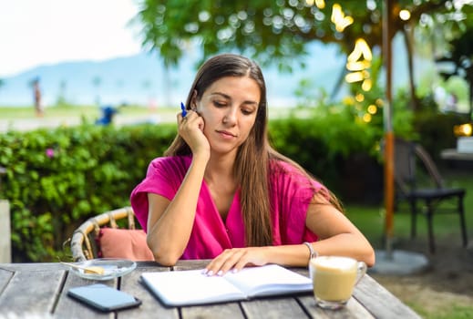 Cute woman sitting on a chair in summer garden and writing in a notebook
