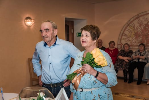 A couple celebrates their 50th wedding anniversary (golden wedding) in the registry office. The wife smiles while holding a bouquet, and their friends are seen in the background, all dressed up