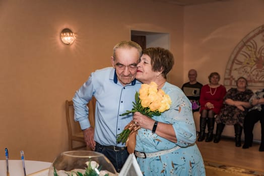 couple celebrates their 50th wedding anniversary (golden wedding) in the registry office. The wife kisses her husband on the cheek while holding a bouquet, and their friends are seen