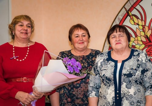 Three Russian friends in festive outfits congratulate their friend on her birthday, holding a bouquet of purple flowers and looking joyful. Warm atmosphere with decorative background