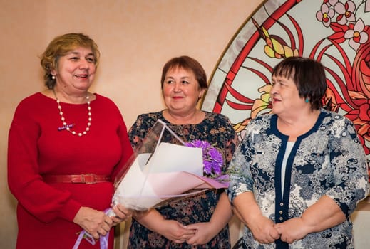Three Russian friends, aged 60 and 70, hold a bouquet of flowers and congratulate their friend on a special occasion. They are looking sideways, dressed in elegant dresses, with a joyful atmosphere