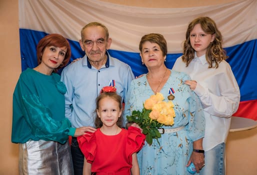 A Russian family celebrates their golden wedding anniversary at the registry office. The elderly couple, over 70, proudly wears medals, with their daughter and granddaughters, and the Russian flag