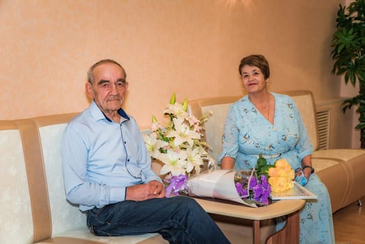 Russian couple over 70 celebrates their golden wedding at the registry office. They sit on sofas in festive attire, smiling at the camera, with bouquets of lilies, roses and chrysanthemums
