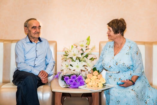Russian couple over 70, celebrating their golden wedding anniversary at the registry office, sit together on sofas. They smile at each other, surrounded by bouquets of lilies, roses, and chrysanthemum