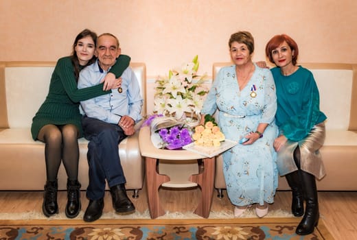 76-year-old man and 71-year-old woman celebrate their golden wedding anniversary, sitting on a sofa in the registry office, embraced by their daughter and granddaughter, holding a bouquet and medal