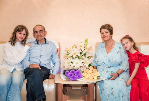 A senior Russian couple just married at the registry office, sitting next to floral bouquets in blue attire. Their granddaughters, one a teenager and the other a young child, hug them