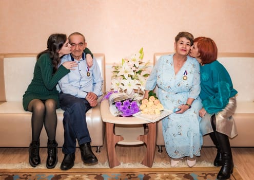 A Russian family celebrating a golden wedding anniversary at a registry office. The husband and wife sit together with medals, surrounded by their daughter and granddaughter