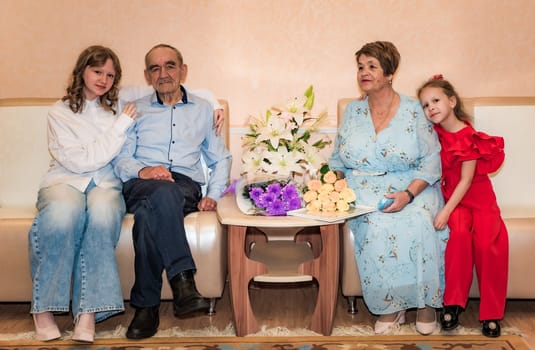 Two granddaughters sit with their Russian grandparents, celebrating their 50th golden wedding anniversary. The family is seated on a sofa, surrounded by bouquets of flowers
