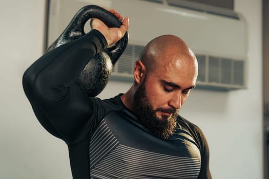 A muscular man with a beard lifts a kettlebell above his head in a workout area. He shows concentration as he engages in strength training, surrounded by gym equipment.