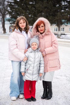 Russian single mother with her teenage and 8-year-old daughters standing in a snowy city. All wear warm winter outfits, including pink and silver jackets. Stylish family winter fashion outdoors