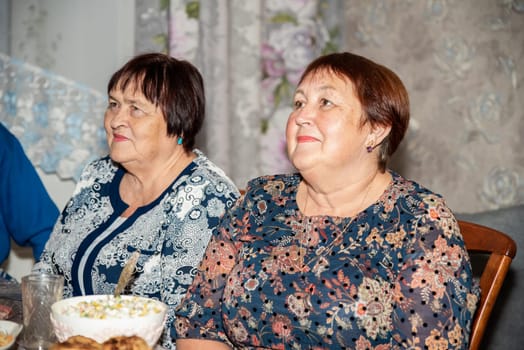 Two Russian women, one 60 and the other 70, look to the side at a television (not visible in the photo). They are dressed in festive attire, with a holiday table and salad in the background