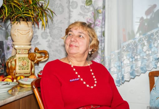 A 60-year-old Russian woman in a red warm dress with beads and short hair sits on a chair at home, looking to the left, enjoying her time as a guest in a cozy indoor setting