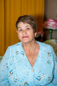 A 71-year-old Russian woman in an elegant festive outfit, featuring a floral-patterned light blue dress. Her short hair, warm smile, and graceful appearance exude charm and dignit