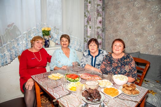 Four Russian friends, two in their 60s and two in their 70s, celebrate a special occasion together. They smile and pose for the camera at the dinner table, enjoying the moment with warmth and joy