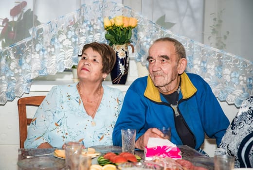 A Russian couple, both over 70 years old, look to the side at a television. They are dressed in festive attire, with glasses on the holiday table in the background