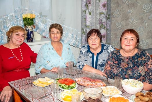 Four Russian women, two in their 60s and two in their 70s, celebrating a festive occasion. The table is filled with traditional