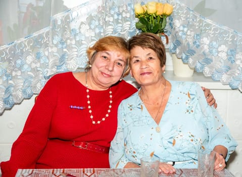 Two cheerful Russian women, aged 60 and 71, smile and embrace at home, celebrating their lifelong friendship. A warm moment of togetherness, perfect for themes of senior friendship and positive aging.