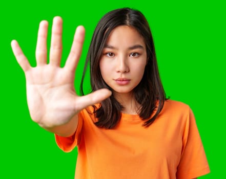 A young woman stands confidently with her hand raised in a stop gesture. She is wearing an orange t-shirt and has dark hair. The vibrant green background enhances the focus on her expression.