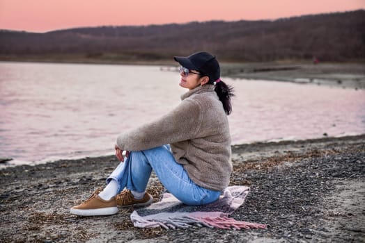 49 year old Russian woman in jacket sitting on a beach and thinking against red sunset