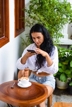 Beautiful ukrainian brunette woman sitting by the table and taking photo of her cappuccino