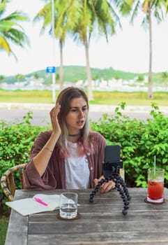 Cute videographer and vlogger taking a video with mobile phone on tripod next to palms and sea. freelance job concept