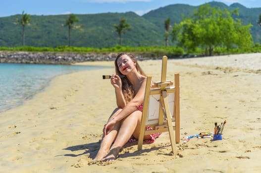 Young artist woman sitting at the beach with a jar with paint brushes and easel close to sea and palms in summer