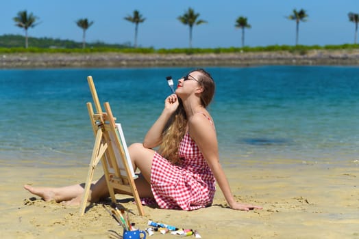 Young artist woman sitting at the beach with a jar with paint brushes and easel close to sea and palms in summer