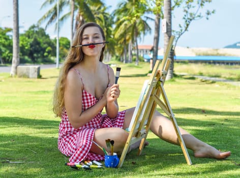 Young artist woman making a funny face with pencil and easel close to sea and palms in summer