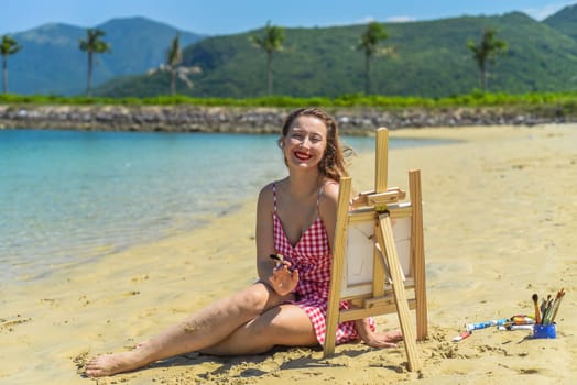 Young artist woman sitting at the beach with a jar with paint brushes and easel close to sea and palms in summer