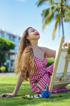 Young artist woman holding a jar with paint brushes and easel close to sea and palms in summer