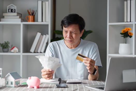A concerned man evaluating his financial situation, holding bills and a credit card, expressing worry in his home office filled with paperwork.