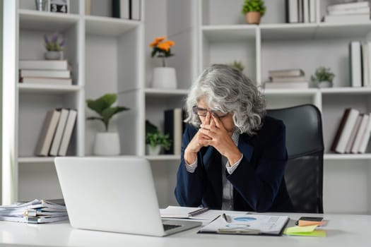 An elderly businesswoman, appearing fatigued, rests her hands on her face while seated at her desk, surrounded by documents and her laptop in a modern office.