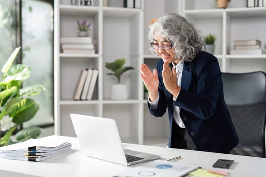 An elderly businesswoman shows her excitement and happiness for a recent accomplishment, interacting with her laptop in a modern office setting.