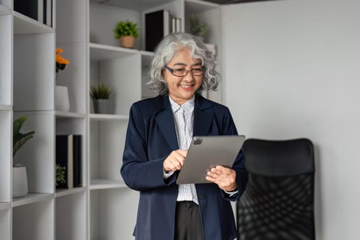 A senior business executive with gray hair happily interacts with a tablet, standing confidently in a contemporary office space filled with plants and organized shelving.